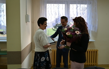widzimy panią stojącą z bukietem kwiat&oacute;w oraz pana przekazującego podarunek drugiej pani