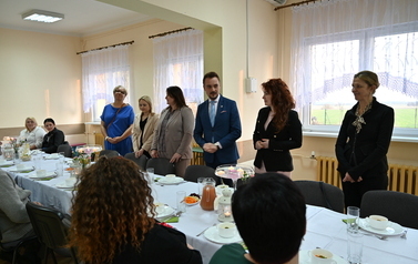 widzimy grupę os&oacute;b siedzących oraz 6 os&oacute;b stojących przy stole 