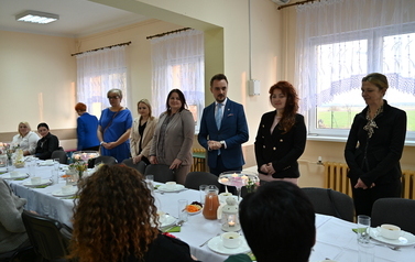 widzimy grupę os&oacute;b siedzących oraz 7 os&oacute;b stojących przy stole 