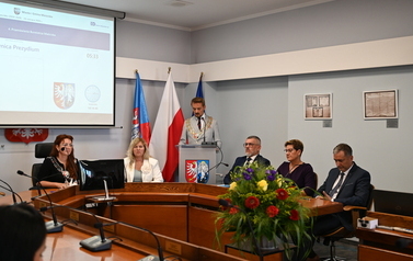 Siedzący radni pośrodku stoi przemawiajacy burmistrz