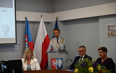 Siedzący radni pośrodku stoi przemawiajacy burmistrz