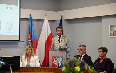 Siedzący radni pośrodku stoi przemawiajacy burmistrz