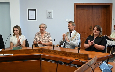 Siedzący radni oraz burmistrz przy stole klacjający w dłonie