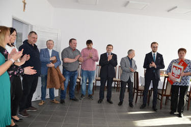 Na zdjęciu widać grupę ludzi kt&oacute;rzy są gośćmi na otwarciu świetlicy. Wśr&oacute;d nich w&oacute;jt gminy - ubrany w granatowy garnitur, pani sołtys w czarnych spodniach i bluzce we wzory. Wszyscy klaszczą.