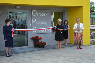 Na zdjęciu widać przemawiającą panią kierownik OPS w Mieścisku. Obok przed wejściem do budynku dwie kobiety trzymają biało-czerwoną wstęgę.