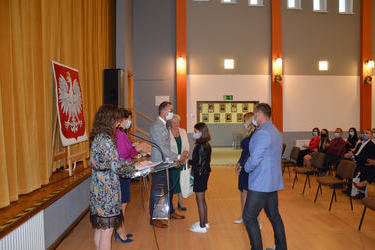 Na zdjęciu w&oacute;jt wręcza stypendium dziewczynce w ciemnych włosach do ramion, ubrana jest w czarną kurtkę i sp&oacute;dnicę. Obok stoją rodzice. Kobieta w jasnych długich włosach, ubrana w granatową sukienkę. Męzczyzna w niebieskiej marynarce i ciemnych spodniach.