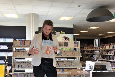 Na zdjęciu widać kobietę trzymającą rysunek. Znajduje się ona w jsnej, nowoczesnej bibliotece.Ubrana jest w ciemną koszulę i ciemne spodnie oraz biały żakiet. Omawia ona trzymaną przez siebie pracę. 