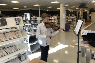 Na zdjęciu widać chłopca grającego na skrzypcach. Znajduje się on w jasnej, nowoczesnej bibliotece. Ubrany jest w białą koszulę i ciemne spodnie.