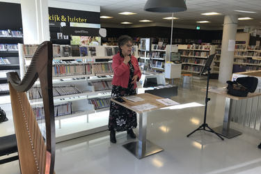 Na zdjęciu znajduje się starsza kobieta stojąca na tle p&oacute;łek z ksiązkami. Znajduje się w jasnej nowoczesnej bibliotece. Ma szare, kr&oacute;tkie włosy, okulary, długą kwiecistą skuknię i czerwony żakiet. Po twarzy widać, że jest w trakcie przemowy.