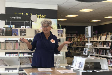 Na zdjęciu znajduje się starsza kobieta stojąca na tle p&oacute;łek z książkami. Znajduje się ona w nowoczesnej bibliotece. Ma siwe kr&oacute;tkie włosy, okulary i granatową bluzkę. Kobieta ma podniesione ręce, gestykuluje podczas powitania gości na wystawie.