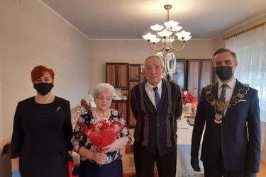 Na zdjęciu znajduje się od lewej strony pani urzędnik w czarnej sukience, następnie starsza pani w odświętnym stroju z kwiatami w ręce, obok niej stoi starszy pan, a obo niego w&oacute;jt w galowym stroju.