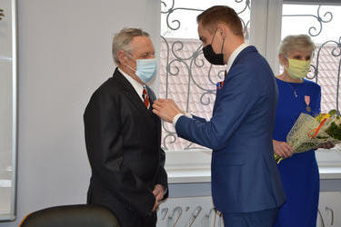 Mężczyzna w granatowym garniturze-W&oacute;jt przypina medal starszemu panu. Mężczyzna ma kr&oacute;tkie siwe włosy, ciemny garnitur.