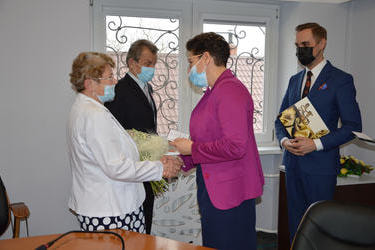 Na zdjęciu przewodnicząca w fuksjowym żakiecie składa gratulacje starszej pani w jasnym żakiecie. Kobieta ma jasne kr&oacute;tkie włosy, w rękach trzyma bukiet kwiat&oacute;w.