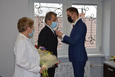 Na zdjęciu W&oacute;jt przypina starszemu mężczyźnie medal. Starszy pan ubrany jest w cimny garnitur. Obok stoi starsza pani ubrana w jasny żakiet, trzyma w rękach kwiaty.