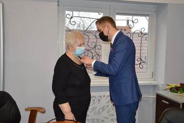 Na zdjęciu znajduje się starsza kobieta wkr&oacute;tkich siwych włosach, ubrana na czarno oraz młody mężczyzna w garniturze-W&oacute;jt, przypina medal starszej pani