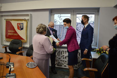Na zdjęciu znajduje się starsza kobieta w r&oacute;żowej garsonce, starszy mężczyzna w ciemnym garniturze, przewodnicząca Rady Gminy w czarnej sukni i fuksjowym żakiecie, ma kr&oacute;tkie ciemne włosy oraz W&oacute;jt Gminy w granatowym garniturze. Młodsza kobieta wręcza 