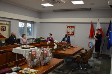 Na zdjęciu znajduje się starsze małżeństwo, przewodnicząca Rady Gminy - kobieta w ciemnych kr&oacute;tkich włosach i fuksjowym żakiecie, W&oacute;jt Gminy - meżczyzna ubrany w granatowy garnitur. Wszyscy siedzą przy dużym stole. Przy m&oacute;wnicy znajduje się kobieta-kierow