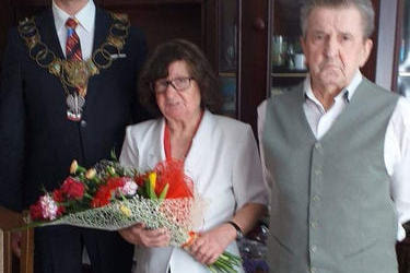 Na zdjęciu znajduje się starsze małżeństwo. Kobieta w ciemnych włosach do ramion, ciemnej sp&oacute;dnicy i białym żakiecie, trzyma w rękach bukiet kwiat&oacute;w. Mężczyzna w białej koszuli, szarych spodaniach i kamizelce. Obok małżeństwa stoi młodszy mężczyzna