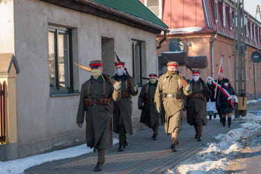 Na zdjęciu znajduje się grupa ludzi przepranych w mundury powstańc&oacute;w z karabinami na plecach. Maszerują ulicą. 
