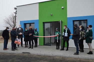 Na zdjęciu widać grupę ludzi stojących koło wejścia do szatni. Budynek w kolorze biało-zielono-niebieskim. Przed wejściem w&oacute;jt Gminy Mieścisko ma przemowę. Ubrany jest w granatowy płaszcz. Obok niego stoją chłopcy w storjach piłkarskich i trzymają wstęgę