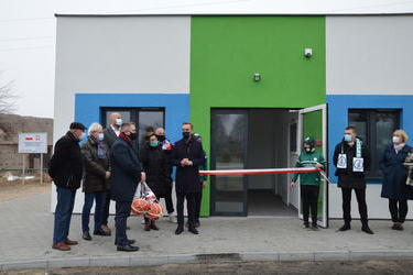 Na zdjęciu widać grupę ludzi stojących koło wejścia do szatni. Budynek w kolorze biało-zielono-niebieskim. Przed wejściem w&oacute;jt Gminy Mieścisko ma przemowę. Ubrany jest w granatowy płaszcz. Obok niego stoją chłopcy w storjach piłkarskich i trzymają biało-c