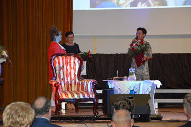 Kobieta wygłasza przemowę w kierunku jubilatki. Trzyma dyplom i duży bukiet kwiat&oacute;w