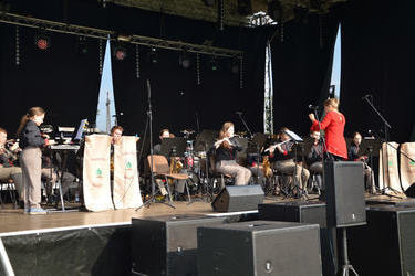 Na zdjęciu widać scenę a na niej orkiestrę dętą wykonującą jakiś utw&oacute;r. Muzykami dyryguje kobieta ubrana w czerwoną tunikę.