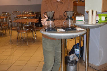 Na zdjęciu widać mężczyznę baristę. Ubrany jest w brązową koszulę i zielone spodnie. Przed nim na stoliczku stoją zaparzacze do kawy.