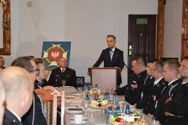 Przy stole siedzą strażacy w mundurach galowych. W tle stoi i przemawia W&oacute;jt.