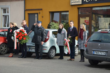 Na zdjęciu widać władze Gminy z wieńcem. W&oacute;jt trzyma wiązanke w kolorze biało-czerwnonym. Obok niego znajduje się przewodnicząca Rady oraz Prezes Gminnego Związku OSP. Obok stoi druga delegacja młodych ludzi - mężczyzna i dwie kobiety.