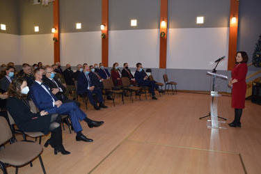 Zdjęcie przedstawia przemawiającą kobietę. Stoi przy m&oacute;wnic. Ubrana jest w czerwoną sukienkę. Widownia patrzy na nią w pełnym skupieniu. 