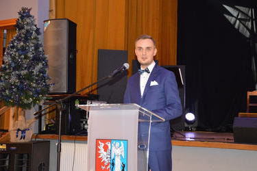 Na zdjęciu widać w&oacute;jta. Stoi przed m&oacute;wincą, wygłasza przem&oacute;wienie. Ubrany jest w jasną koszulę i granatow garnitur.