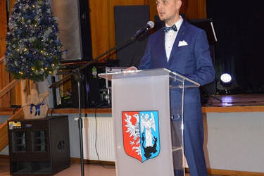 Na zdjęciu widać w&oacute;jta. Stoi przed m&oacute;wincą, wygłasza przem&oacute;wienie. Ubrany jest w jasną koszulę i granatow garnitur.