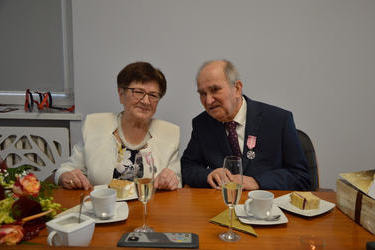 Na zdjęciu widać starsze małżeństwo. Kobieta w kr&oacute;tkich , ciemnych włosach, ma okulary, ubrana w kolorową sukienkę i jasny żakiet. Mężczyzna w jasnej koszuli i ciemnym garniturze. Oboje mają przypięte medale.