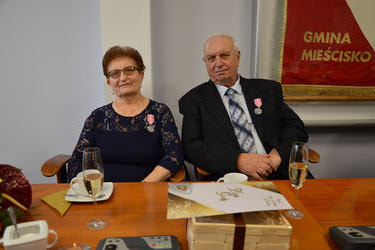 Na zdjęciu widać starsze małżeństwo. Kobieta w kr&oacute;tkich włosach, ma okulary, ubrana w granatową sukienkę. Mężczyzna w siwych włosach, w ciemnym garniturze i białej koszuli. Obobje na piersi mają medale.