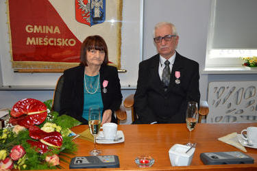 Na zdjęciu widać starsze małżeństwo. Kobieta w ciemnych włosach do ramion, ubrana w zieloną sukienkę i ciemny żakiet. Mężczyzna w siwych włosach, okularach,w ciemnym garniturze. Oboje na piersi mają medale.