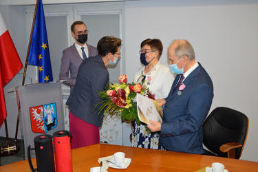 Na zdjęciu w&oacute;jt wraz z przewodniczącą rady wręczają parze jubilat&oacute;w medal, gratulacje i kwiaty. Starsi ludzie są ubrani elegancko. Kobieta w sukience i jasnym żakiecie. Mężczyzna w garnatowym garniturze.