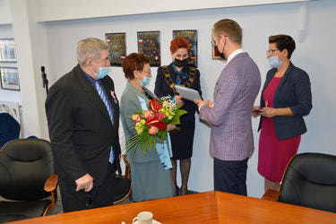 Na zdjęciu w&oacute;jt wraz z przeowdniczącą rady i kierownikiem USC wręczają jubilatom gratulacje, medale i kwiaty. Starsze małżeństwo elegancko ubrane.
