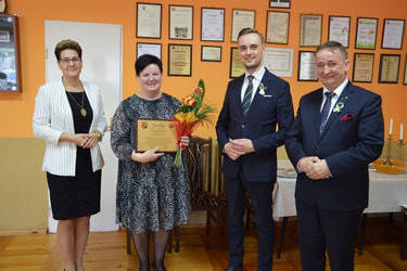 Zdjęcie przedstawia od lewej strony przeowdniczącą Rady Gminy ubraną w czarną sukienkę i biały żakiet. Obok stoi pani Sołtys obchodząca jubileusz, trzyma w ręce grawerton i kwiaty. Następnie jest w&oacute;jt Gminy Mieścisko w granatowym garniturze. 