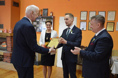 W&oacute;jt wręcza grawerton sołtysowi kt&oacute;ry obchodzi jubileusz 30lecia bycia sołtysem. Oboje mężczyzn ubranych jest w ciemne garnitury. Obok w&oacute;jta stoją goście.
