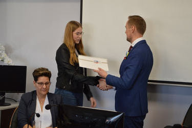 Na zdjęciu w&oacute;jt wręcza dokumenty i gratulacje młodej radnej. Jest to dziewczyna w długich, jasnych włosach, ma okulary. Ubrana jest w jasna bluzkę i ciemną marynarkę.