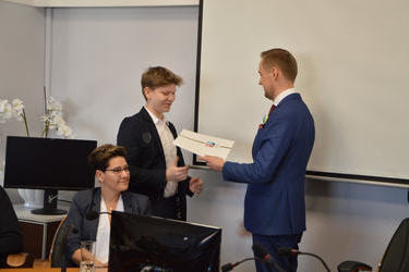 Na zdjęciu w&oacute;jt wręcza dokumenty i gratulacje młodemu radnemu. Jest to chłopak w kr&oacute;tkich, blond włosach. Ubrany w białą koszulę i ciemna marynarkę.