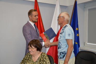 W&oacute;jt Gminy Mieścisko podaje rękę mężczyznie z Holandii wręczając mu upominek na tle flag. Przed nimi siedzi kobieta.