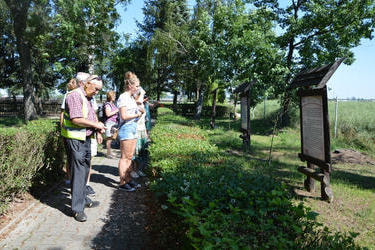 Goście na świażym powietrzu,czytają tablicę informacyjną. Dookoła nich zielone krzewy i drzewa. Na pierwszym planie widać pana, kt&oacute;ry ma ubraną kamizelkę odblaskową.