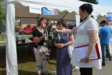 Trzy kobiety stoją przed namiotem. Jedna z nich trzyma swoją konstrukcje kwiatową wykonaną na patyku, obok kobieta o niej opowiada. Prowadząca trzyma jej mikrofon.
