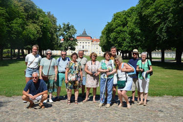 Zdjęcie grupowe gości z Francji i Holandii przed zamkiem. Jeden pan kuca. Po obu stronach rosną drzewa.