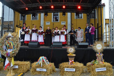 Zdjęcie przedstawia zesp&oacute;ł ludowy Sarbianie występujący na scenie. Obok stoi w&oacute;jt z przewodniczącą rady. Przed sceną znajdują się wieńce dożynkowe.
