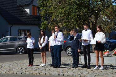 Zdjęcie przedstawia młodzież w eleganckich ubraniach. Jeden z chłopak&oacute;w trzyma w ręce mikrofon i recytuje wiersz.