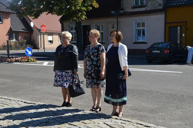 Zdjęcie przedstawia trzy kobiety stojące przed pomnikiem. Są one elegancko ubrane. Dwie mają suknie w kwiaty, trzecia granatową i biały żakiet.