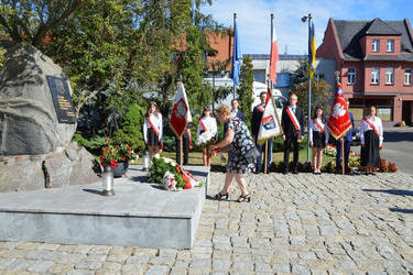 Na zdjęciu widać kobietę,kt&oacute;ra składa kwiaty na pomniku. Ubrana jest w kwiecistą sukienkę. W tle znajdują się delegacje ze sztandarami.